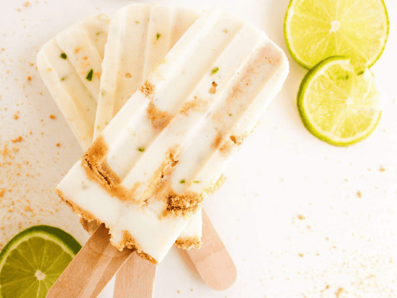 Three creamy popsicles with lime slices and crumbs scattered around on a white surface.