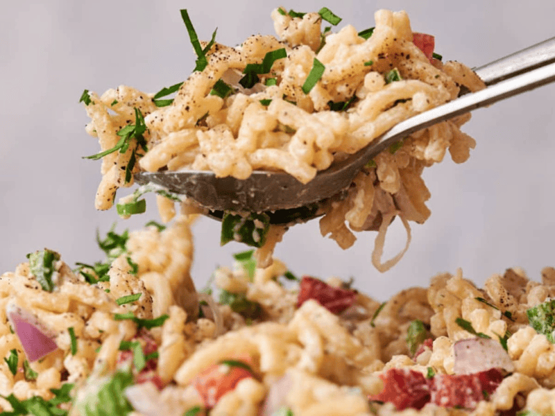 A spoon lifting a serving of creamy macaroni salad with diced red onion and fresh herbs, with pepper sprinkled on top.