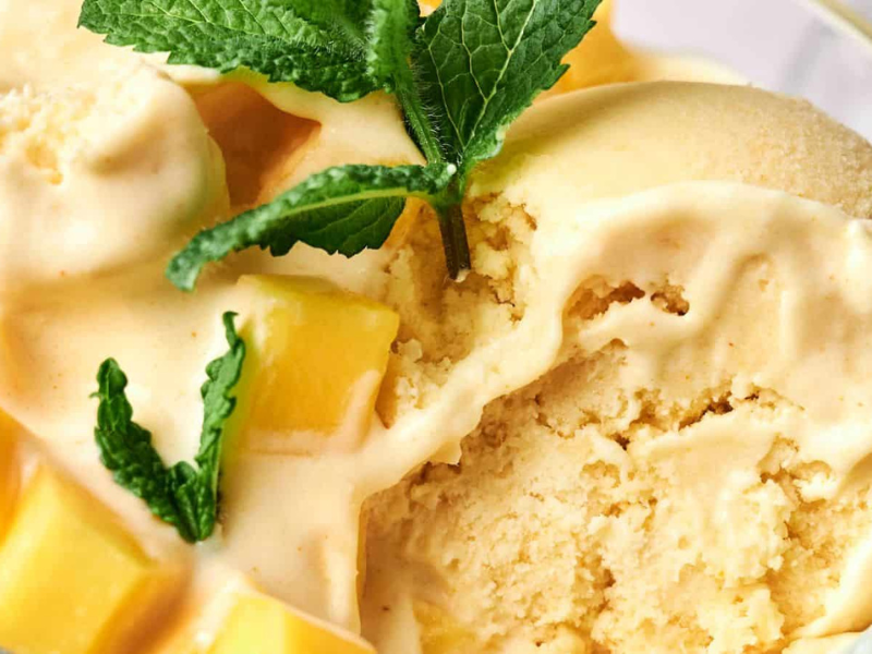 A bowl of creamy mango ice cream is garnished with fresh mango cubes and mint leaves.