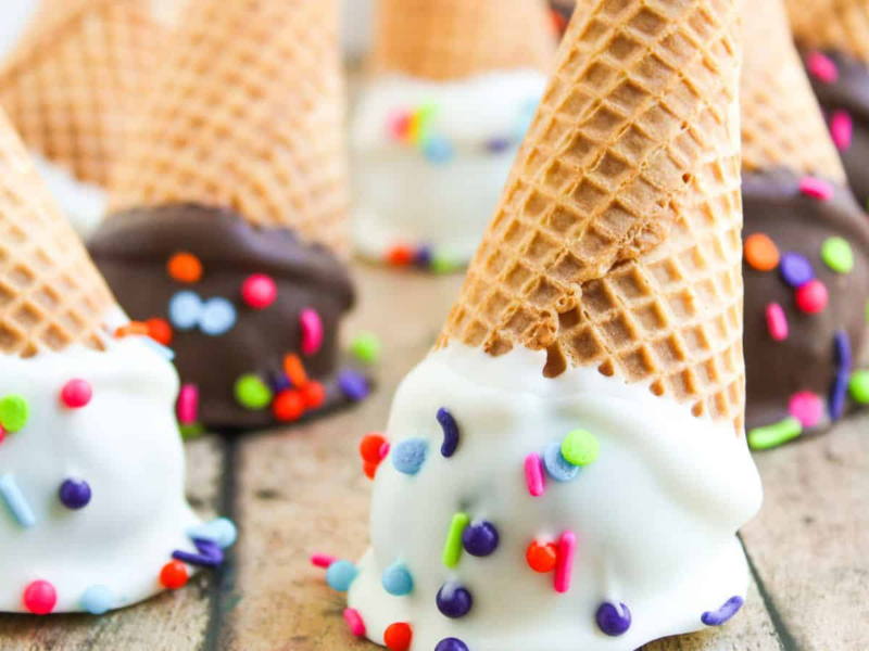 Waffle ice cream cones dipped in white and dark chocolate, decorated with colorful sprinkles, arranged on a wooden surface.