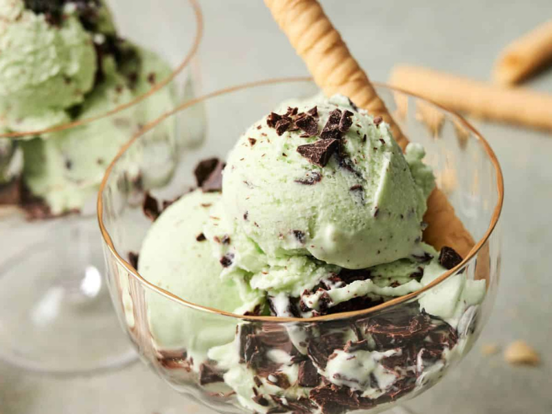 A glass dessert cup filled with mint chocolate chip ice cream, topped with chocolate pieces and served with rolled wafer cookies.