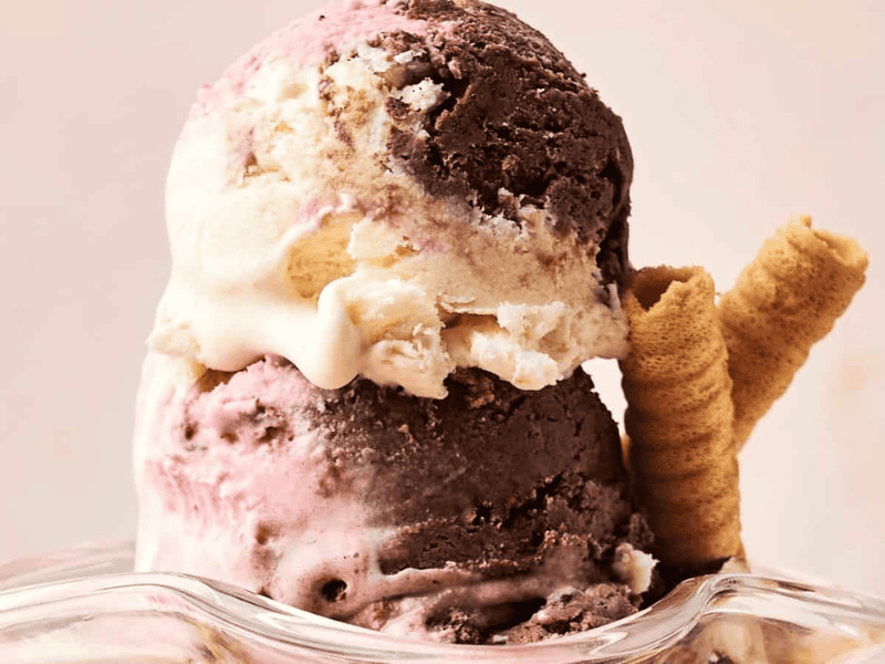 Two scoops of Neapolitan ice cream in a glass dish, featuring chocolate, vanilla, and strawberry flavors, served with two rolled wafer cookies.
