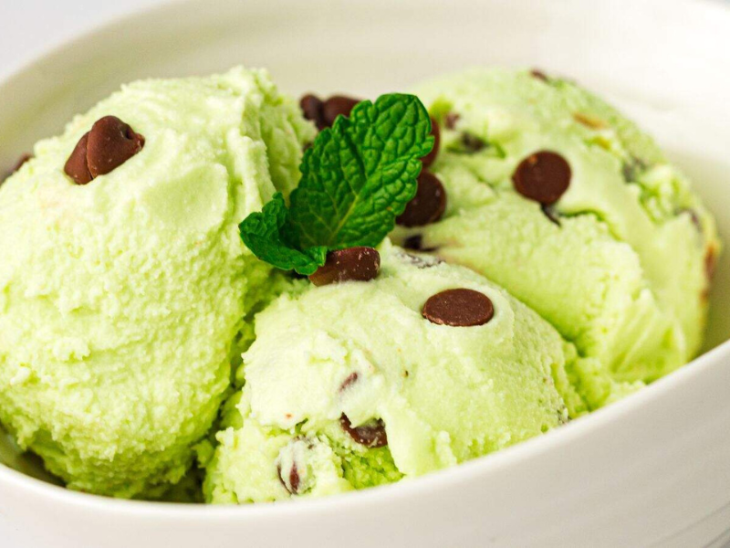 Three scoops of mint chocolate chip ice cream with chocolate chips, garnished with a sprig of mint, in a white bowl.