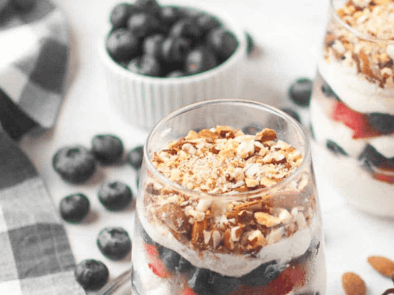 A glass filled with layers of cheesecake and berries sits on a table, surrounded by scattered blueberries and a bowl of blueberries in the background.