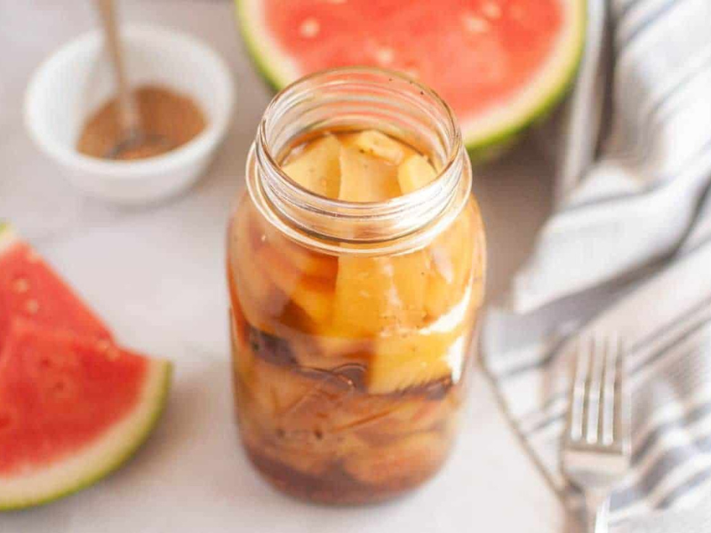 A jar of pickled watermelon rind sits on a white surface, with watermelon slices, a striped cloth, a fork, and a small bowl of spices in the background.