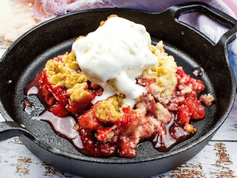 A cast iron skillet with strawberry rhubarb cobbler topped with a scoop of vanilla ice cream on a wooden surface with a cloth napkin beside it.