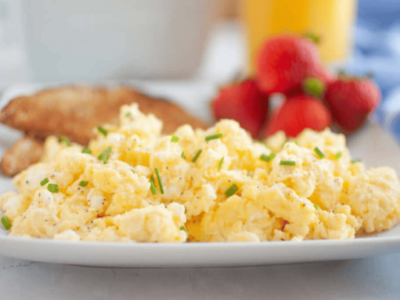 A plate of scrambled eggs garnished with chives, served with toast and strawberries, with a glass of orange juice in the background.