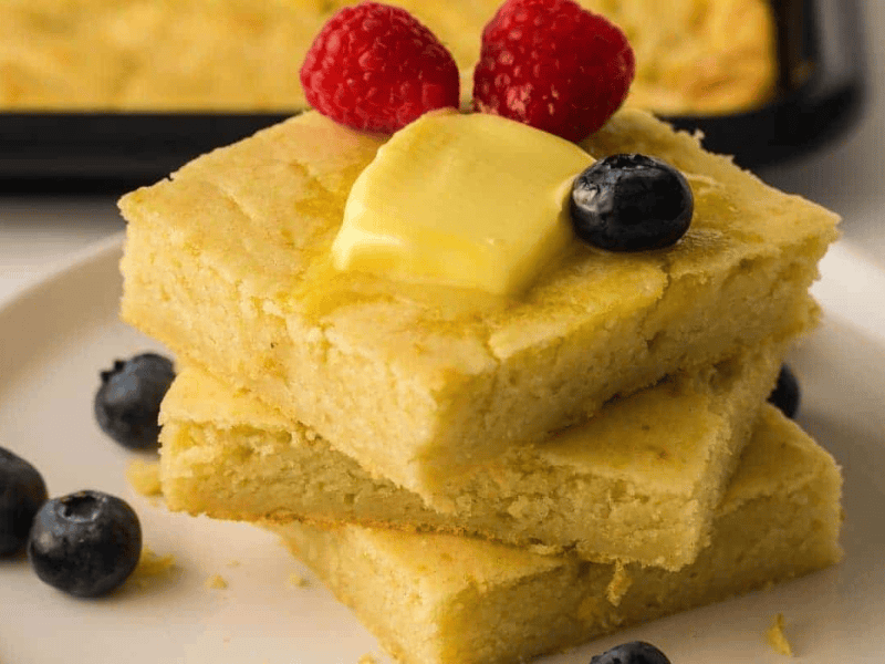 Three stacked pieces of cornbread topped with a pat of butter, raspberries, and a blueberry, with additional blueberries on the plate.