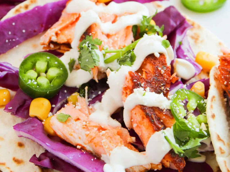 Tacos with grilled salmon, purple cabbage, corn, jalape&ntilde;o slices, cilantro, and drizzled with white sauce on tortillas.