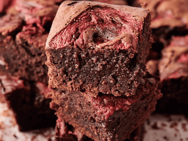 Two pieces of chocolate brownies with a swirl of red fruit, possibly raspberry or cherry, stacked on a surface with more brownies in the background.