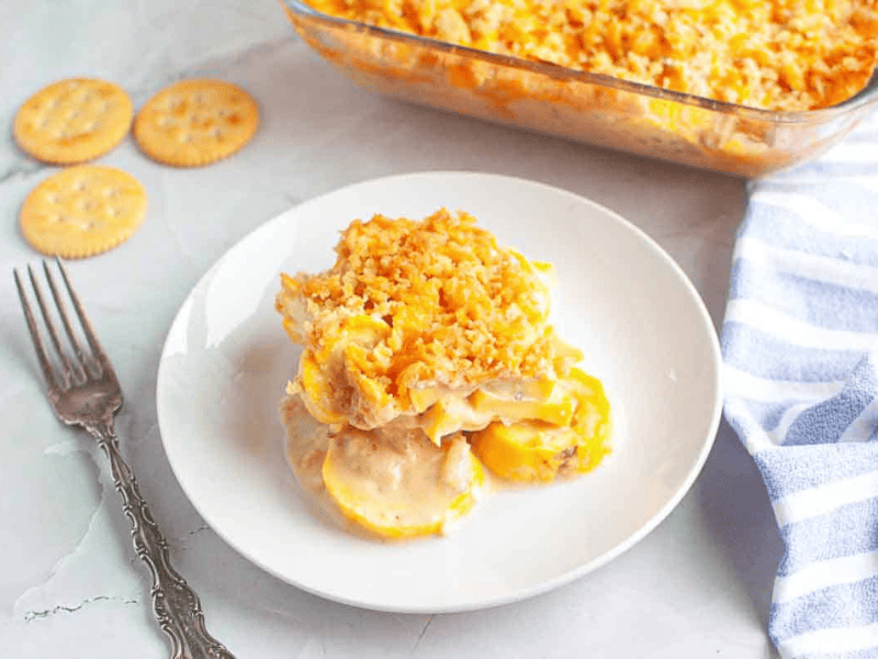 A serving of yellow squash casserole with a crispy crumb topping on a white plate, next to a fork, crackers, and a glass baking dish.