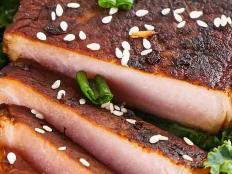 Close-up of sliced grilled pork belly garnished with sesame seeds and green chili, served on a bed of kale.