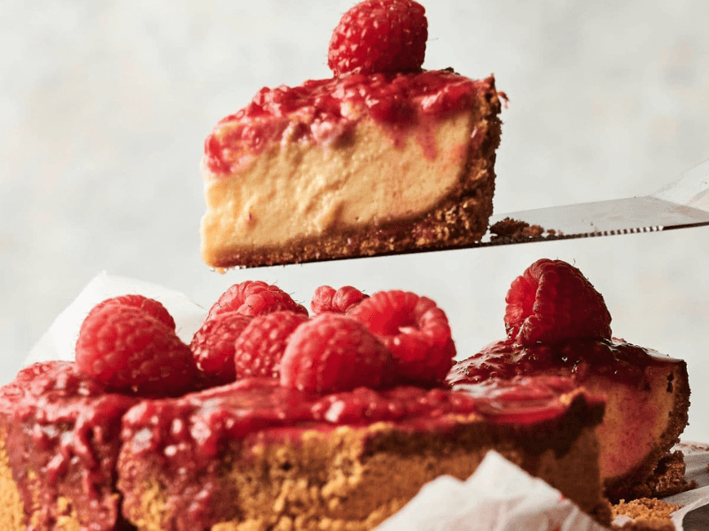 A slice of raspberry-topped cheesecake is being lifted from a whole cheesecake with a graham cracker crust.