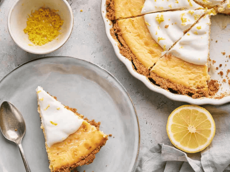 A slice of lemon pie sits on a plate next to a pie with a slice removed.