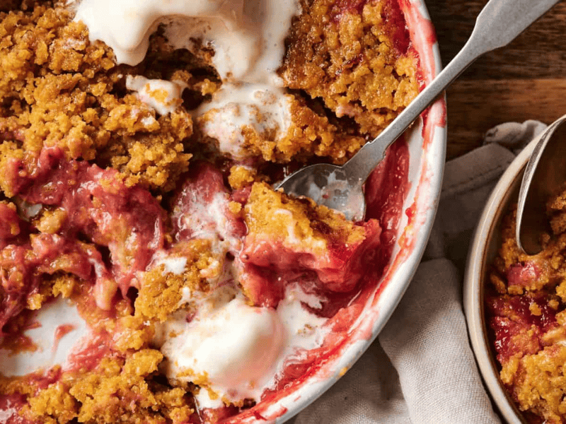 A baked dessert with a crumbly brown topping and red filling sits in a dish, with a portion served in a separate bowl. Spoons are present in both the dish and the bowl.