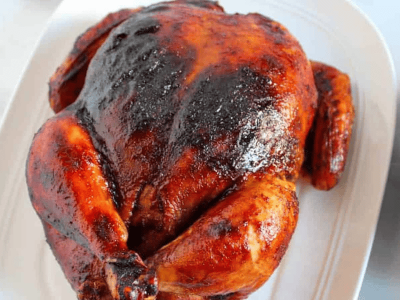 Roasted chicken with a crispy, golden-brown skin on a white plate, served with a side of greens in a black bowl.