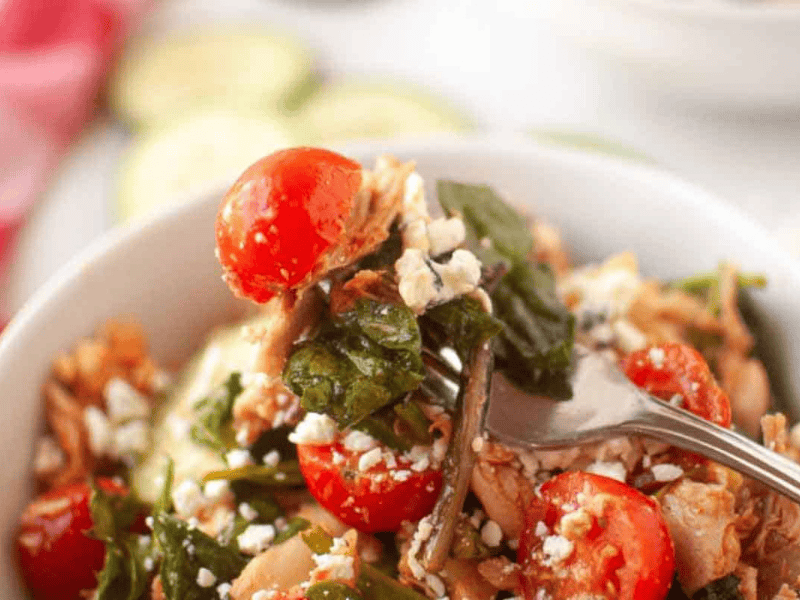 A close-up of a fork in a bowl of salad with cherry tomatoes, leafy greens, shredded chicken, and crumbled cheese.