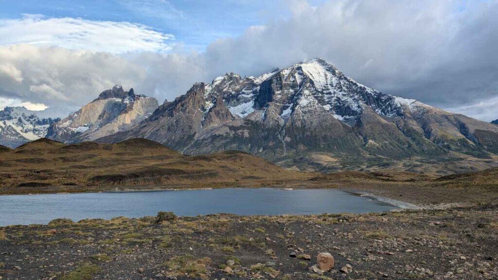 Snow-capped mountains rise behind a small lake and rolling hills under a partly cloudy sky.