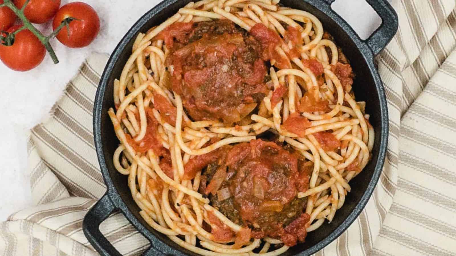 A cast iron skillet filled with spaghetti and tomato sauce, topped with three large meatballs. A striped towel and vine tomatoes are nearby.