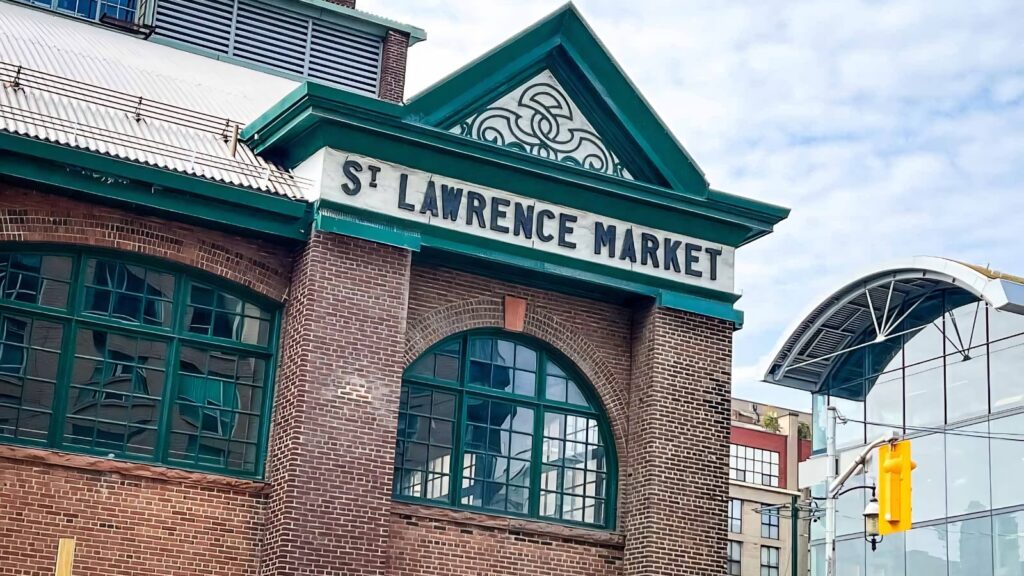 St. Lawrence Market building with green trim, large arched windows, and a sign on the brick facade; modern glass structure visible on the right.