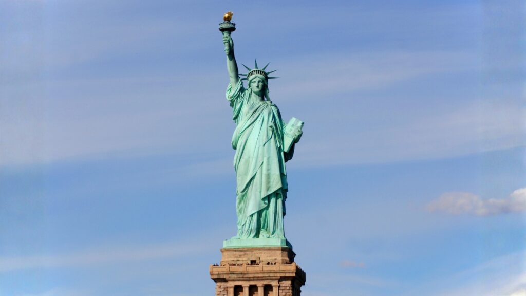 The Statue of Liberty stands on its pedestal against a blue sky, holding a torch in one hand and a tablet in the other.