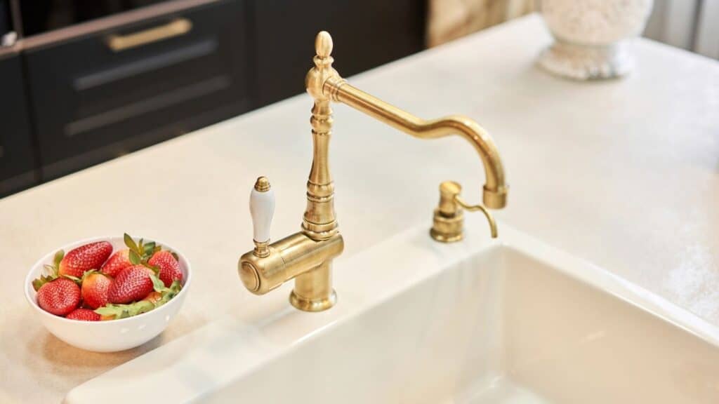 A brass kitchen faucet is installed on a white countertop next to a white bowl of strawberries.