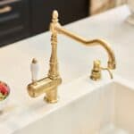 A brass kitchen faucet is installed on a white countertop next to a white bowl of strawberries.