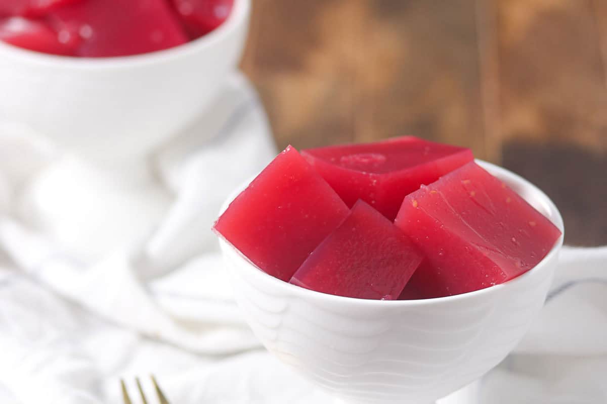 A white bowl filled with red gelatin cubes sits on a white cloth, with another bowl of gelatin in the background.