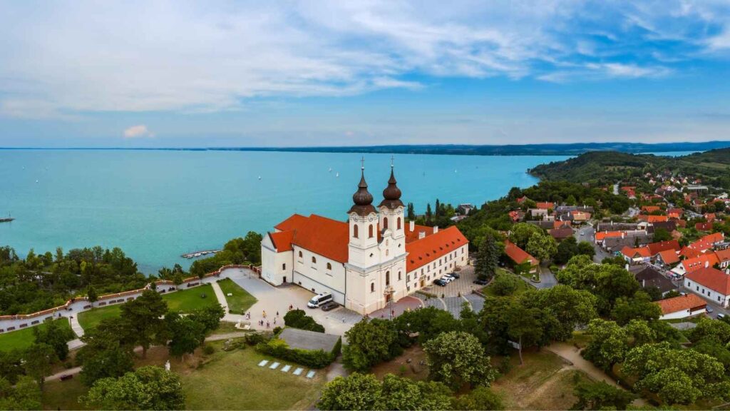 Aerial view of Tihany Abbey with its twin towers, red roof, and surrounding buildings next to Lake Balaton, with green trees and a village nearby.