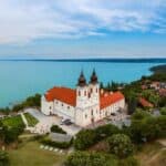 Aerial view of Tihany Abbey with its twin towers, red roof, and surrounding buildings next to Lake Balaton, with green trees and a village nearby.