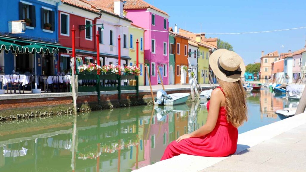 A woman in a red dress and straw hat sits beside a canal lined with colorful buildings and boats on a sunny day.