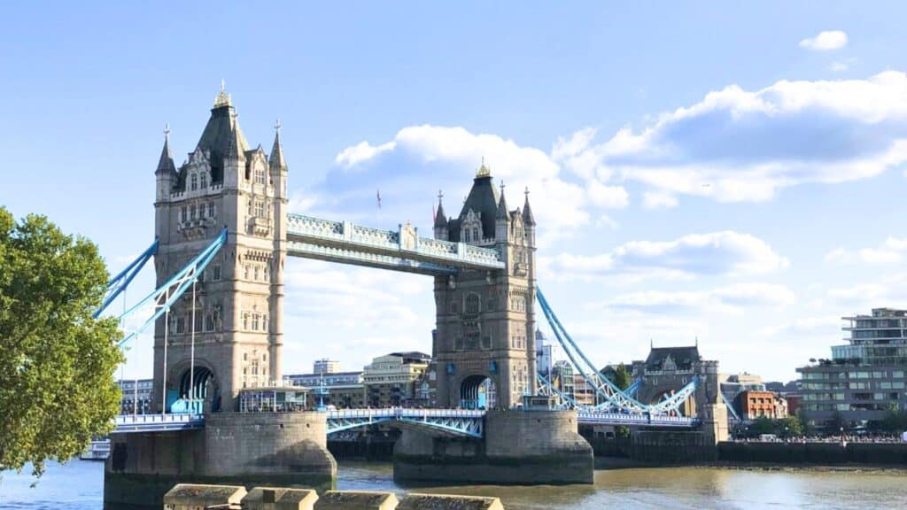 Tower Bridge in London spans the River Thames under a blue sky with scattered clouds, with buildings and trees visible on both riverbanks.