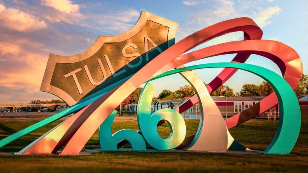 Colorful Route 66 sculpture with the word "Tulsa" stands on grass under a partly cloudy sky, celebrating Route 66 in Tulsa, with trees and a building in the background.