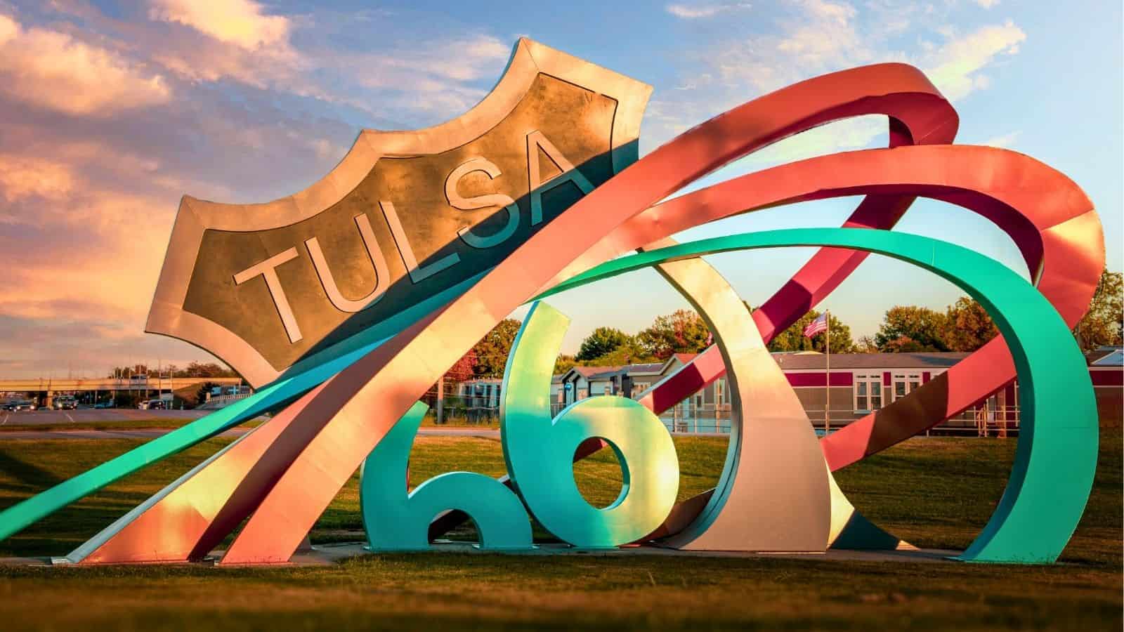 Colorful Route 66 sculpture with the word "Tulsa" stands on grass under a partly cloudy sky, celebrating Route 66 in Tulsa, with trees and a building in the background.