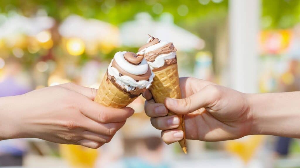 Two hands holding and clinking soft serve ice cream cones with chocolate and vanilla swirls outdoors on a sunny day.