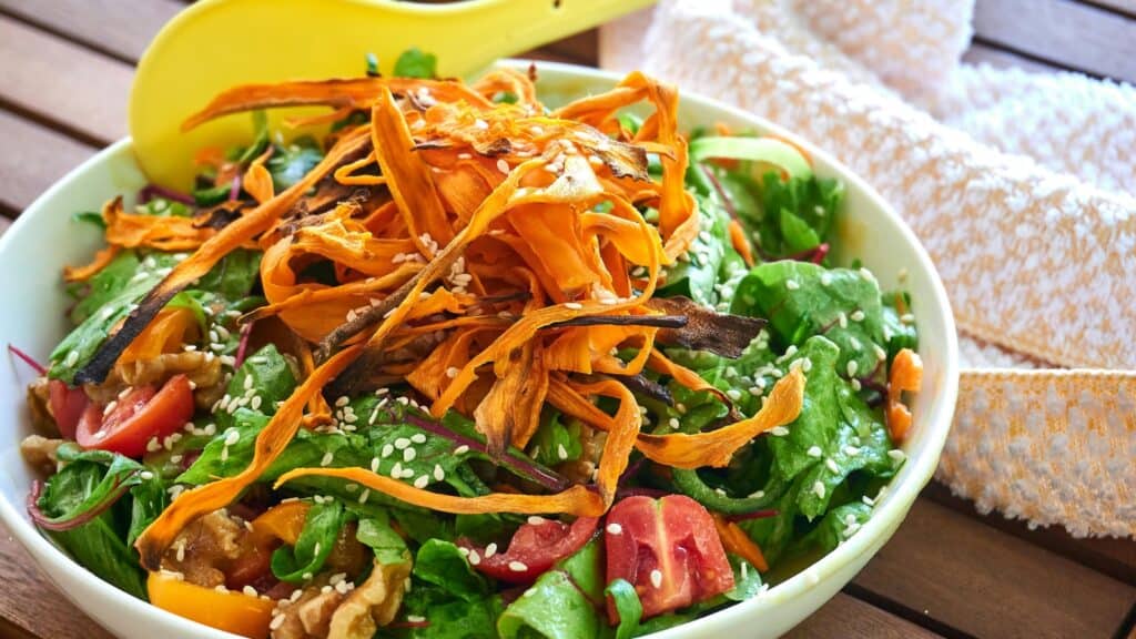 A bowl of fresh salad with cherry tomatoes, greens, walnuts, sesame seeds, and thin crispy carrot strips sits beside a yellow spoon and a textured white cloth&mdash;perfect for celebrating National Salad Month.