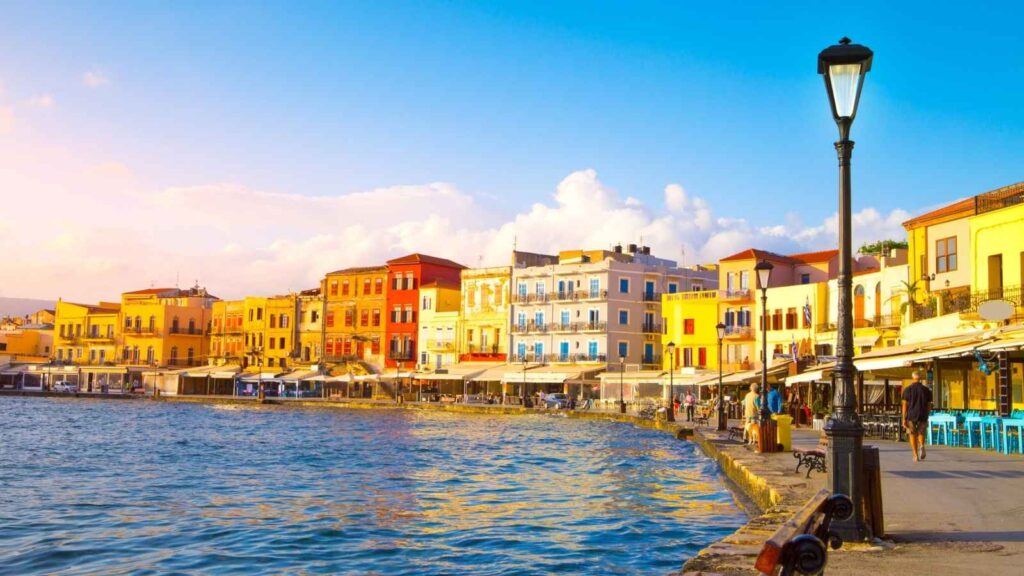 A waterfront promenade in one of the charming European destinations, lined with colorful buildings, outdoor cafes, and a streetlamp beside calm blue water under a clear sky.