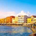 A waterfront promenade in one of the charming European destinations, lined with colorful buildings, outdoor cafes, and a streetlamp beside calm blue water under a clear sky.