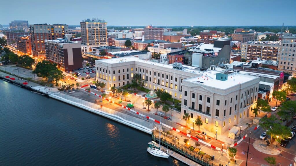 Aerial view of downtown buildings and waterfront area at dusk, with city lights reflecting along the river and a docked boat in the foreground.