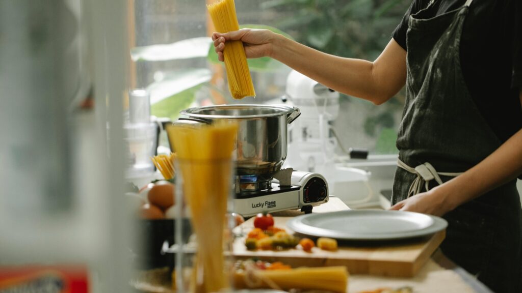 Person holding uncooked spaghetti over a pot on a stove during a pantry challenge, preparing to cook. Various ingredients and kitchen tools are on the counter nearby.