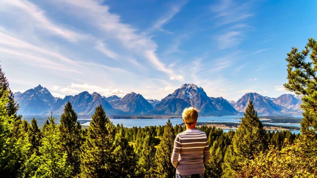 A person with short blond hair stands among pine trees, gazing at distant mountains and a lake under a blue sky with wispy clouds&mdash;a scene straight from a Jackson Hole travel guide.