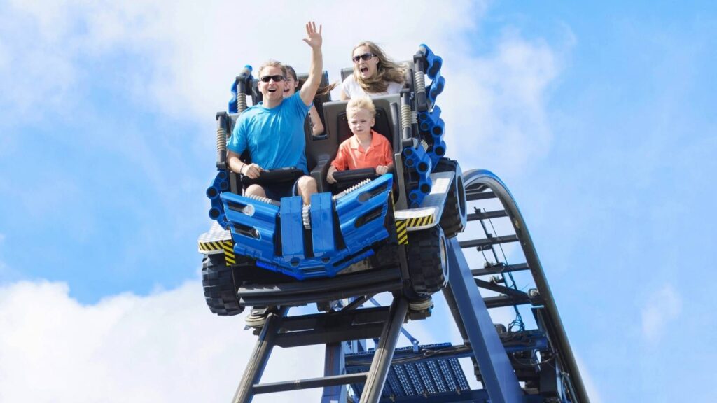 Three people ride a blue roller coaster on a sunny day; two adults in back are excited, while a child in front looks serious&mdash;capturing the thrill and variety of emotions on family vacations.