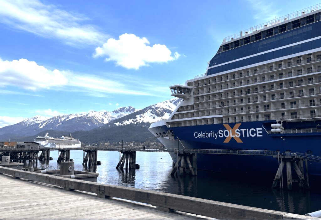 Two cruise ships are docked at a pier with snow-capped mountains and blue sky in the background. The nearest ship, Celebrity Solstice, stands out against the stunning scenery.