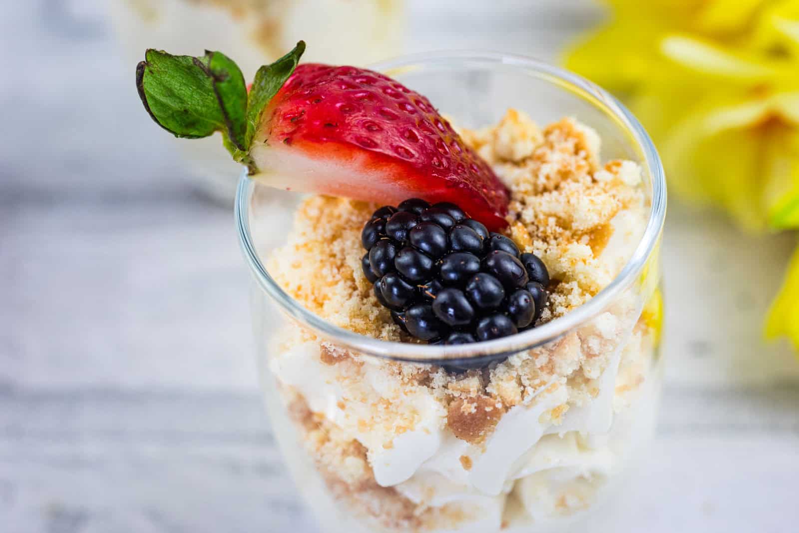 A dessert cup filled with crumbled cookies, cream, topped with a sliced strawberry and a blackberry, set on a light surface with yellow flowers in the background.