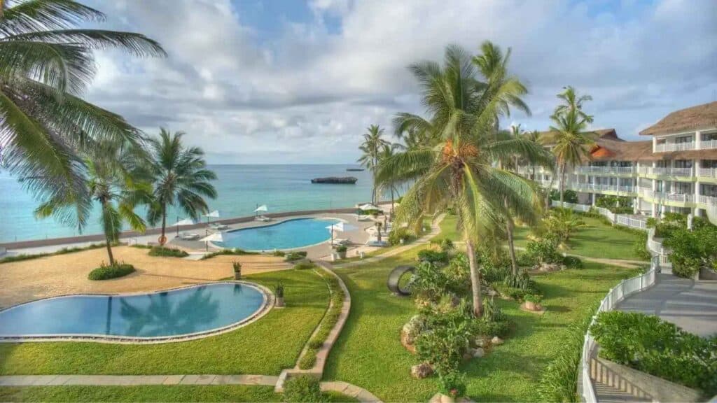 A beachfront resort with palm trees, swimming pools, lounge chairs, and a view of the ocean under a partly cloudy sky.