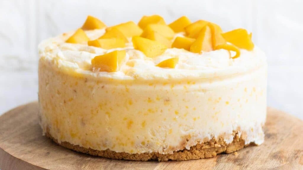A round cheesecake with a crumbly crust, topped with diced mango pieces, sits on a wooden board.