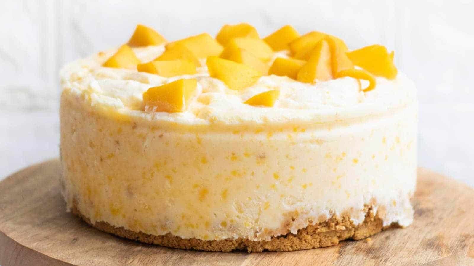 A slice of creamy cheesecake with a crumbly crust, topped with chunks of fresh mango, sits on a white plate with a fork.