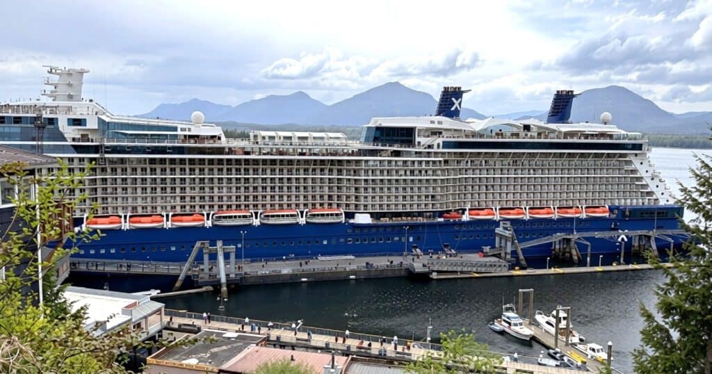 A large Alaska cruise ship docked at a pier with lifeboats visible on its side; small boats and buildings are in the foreground, with mountains in the background.