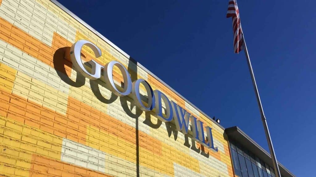 Exterior of a Goodwill store with a large sign on a yellow and orange brick wall, and an American flag on a flagpole against a clear blue sky.