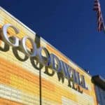 Exterior of a Goodwill store with a large sign on a yellow and orange brick wall, and an American flag on a flagpole against a clear blue sky.
